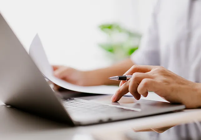 A person typing on a laptop while referencing a document.