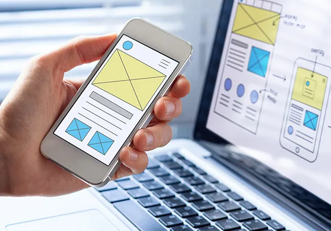 A person holding a phone next to a laptop with web design features displayed on both screens.