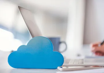 A person writing on a laptop with a cloud next to the laptop to represent cloud computing.