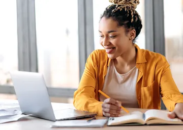 A woman writing in a notebook looking at her laptop screen.