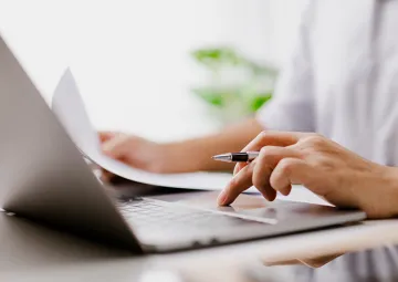 A person typing on a laptop while referencing a document.